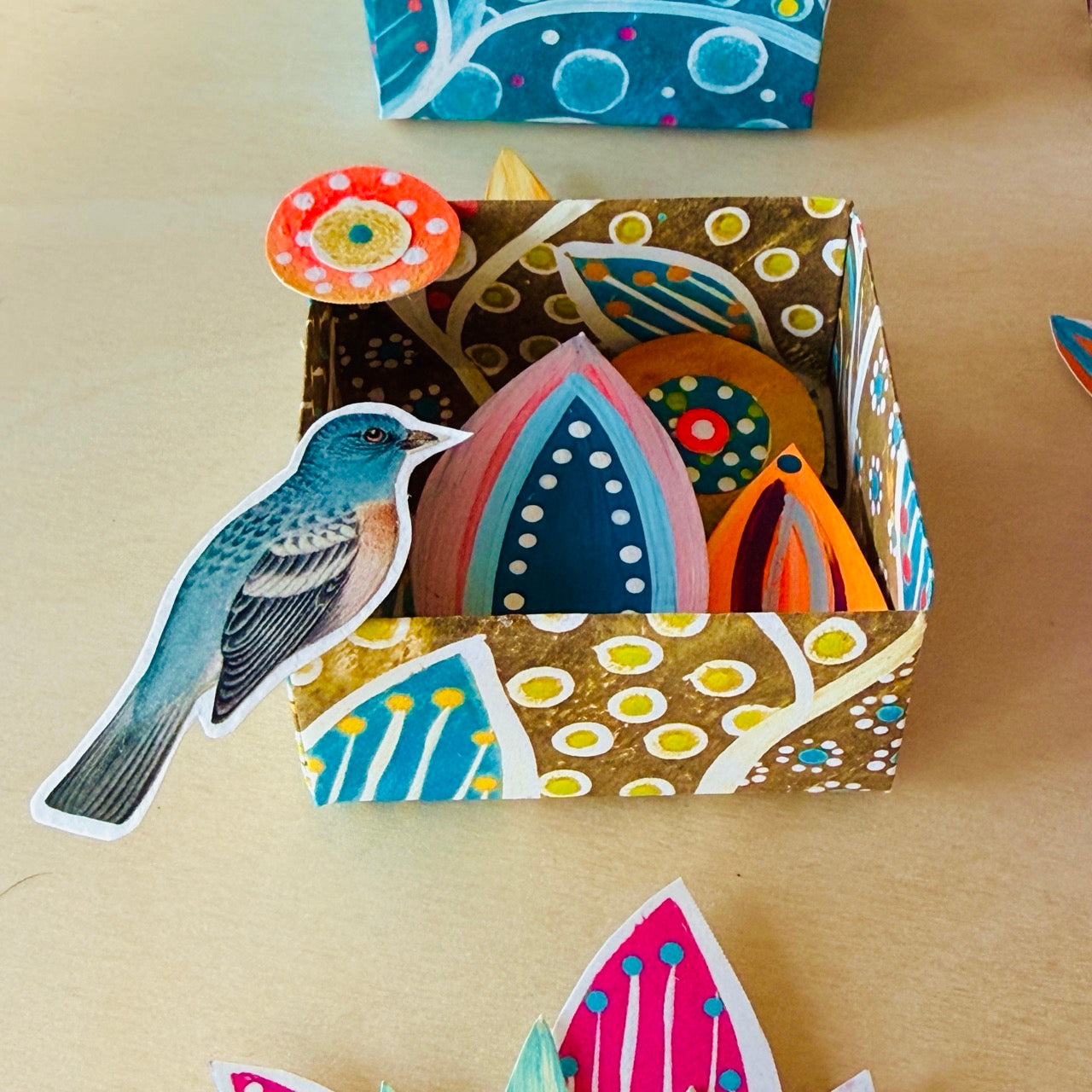 Colorful paper cutouts of birds and flowers in a patterned box on a beige surface.