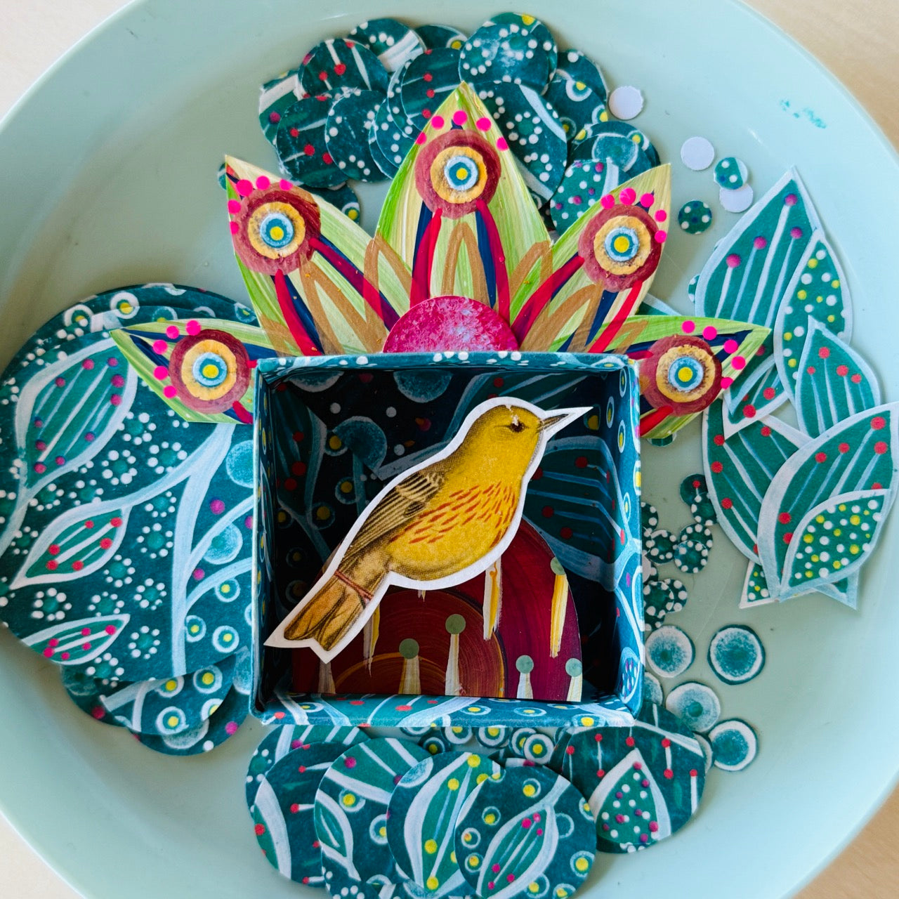Decorative box with a bird illustration surrounded by colorful paper leaves on a white plate.