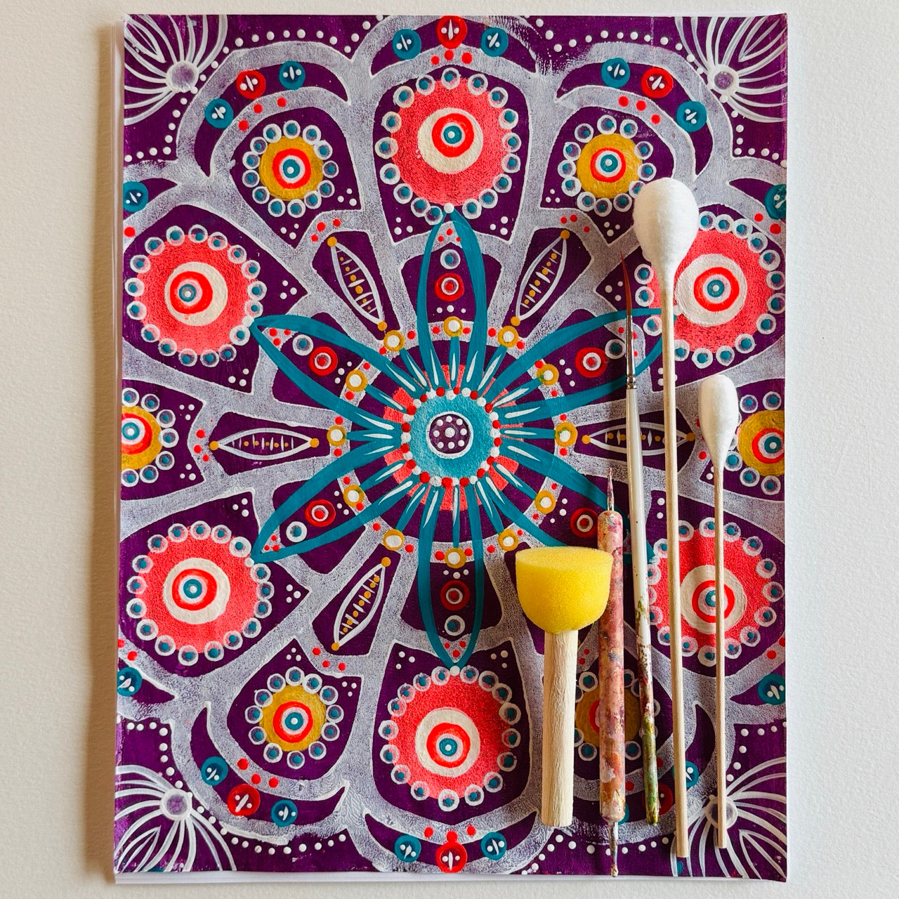 Original hand-painted mandala-inspired artwork featuring layered floral and geometric motifs in vibrant purples, teals, coral, and gold, shown with paintbrushes and tools alongside the canvas.